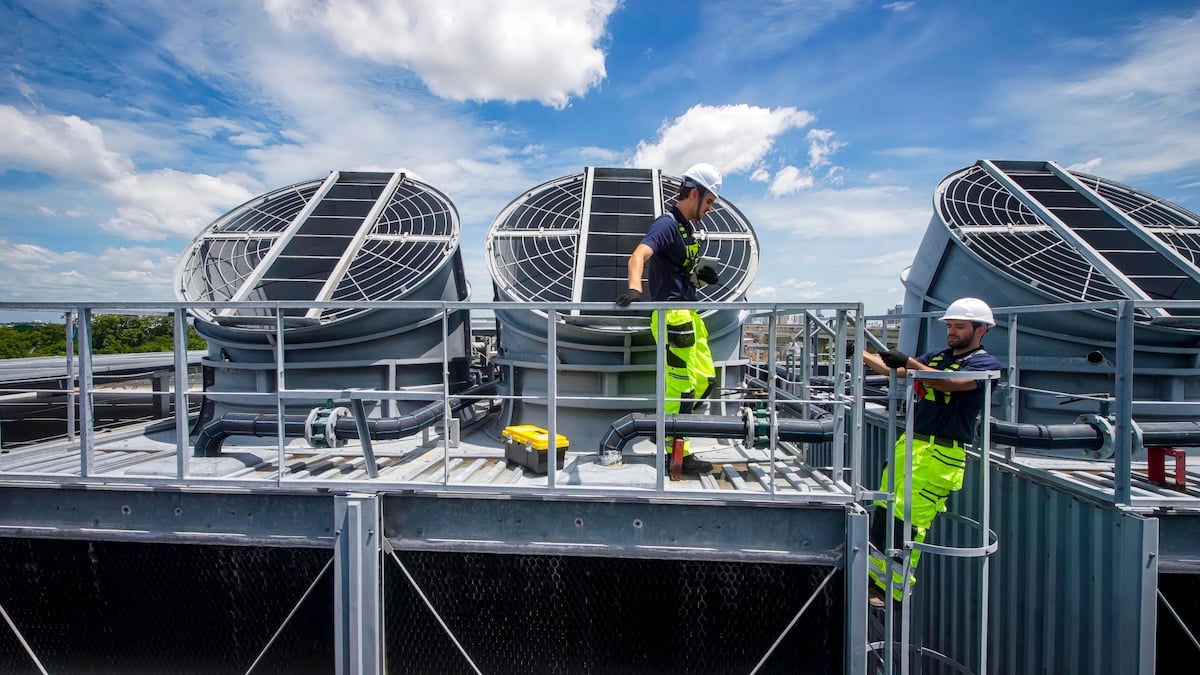 Two skilled workers perform maintenance on large cooling towers under a clear blue sky.